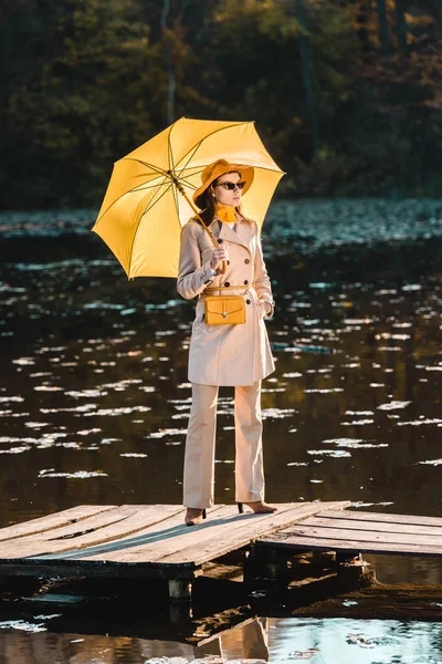 Selective focus of fashionable woman in sunglasses, trench coat and hat posing with yellow umbrella near pond in park — Stock Photo