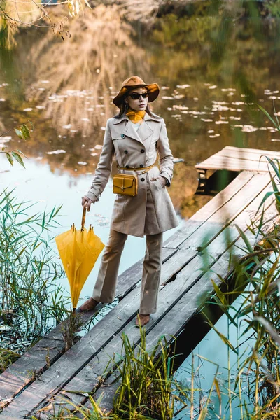 Стильна жіноча модель в сонцезахисних окулярах, тренч пальто і капелюх позує з жовтою парасолькою біля ставка в парку — стокове фото