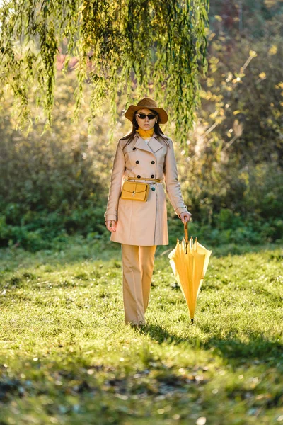 Elegante junge Frau mit Sonnenbrille, Trenchcoat und Hut posiert mit gelbem Regenschirm auf der Wiese — Stockfoto