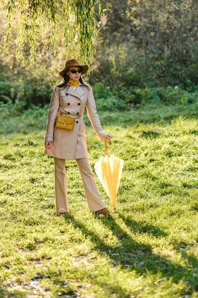 Modische Frau mit Sonnenbrille, Trenchcoat und Hut posiert mit gelbem Regenschirm auf Wiese — Stockfoto