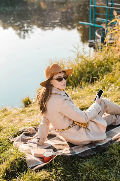 Bela mulher elegante em chapéu e óculos de sol segurando smartphone e sentado em cobertor perto lagoa no parque — Fotografia de Stock