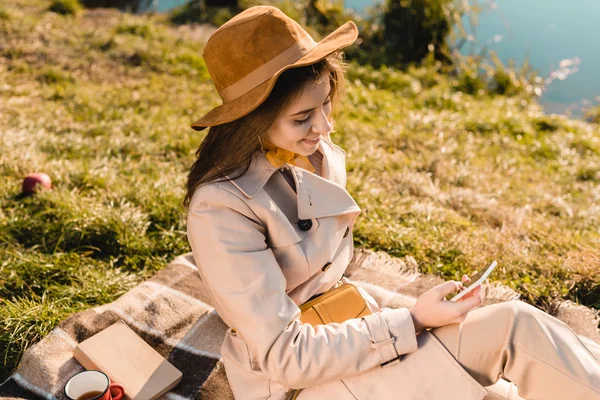 Hochwinkelaufnahme einer lächelnden modischen Frau mit Hut, die ihr Smartphone benutzt, während sie auf einer Decke im Freien sitzt — Stockfoto