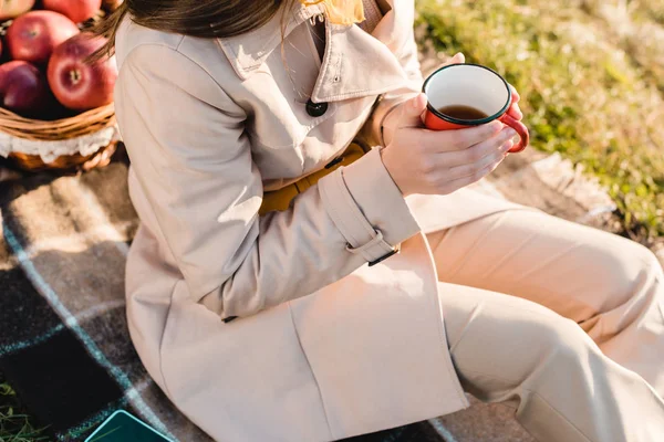 Cropped image of fashionable young woman in trench coat holding cup of coffee while sitting on blanket outdoors — Stock Photo