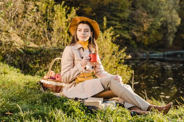 Selective focus of stylish young woman in trench coat and hat holding cup of coffee while sitting on blanket outdoors — Stock Photo