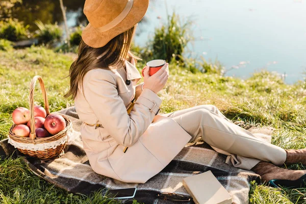 Vista lateral da jovem mulher em casaco de trincheira e chapéu segurando xícara de café enquanto sentado em cobertor ao ar livre — Fotografia de Stock