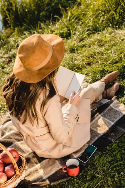 Visão traseira da mulher elegante elegante no livro de leitura de chapéu no cobertor perto da lagoa ao ar livre — Fotografia de Stock