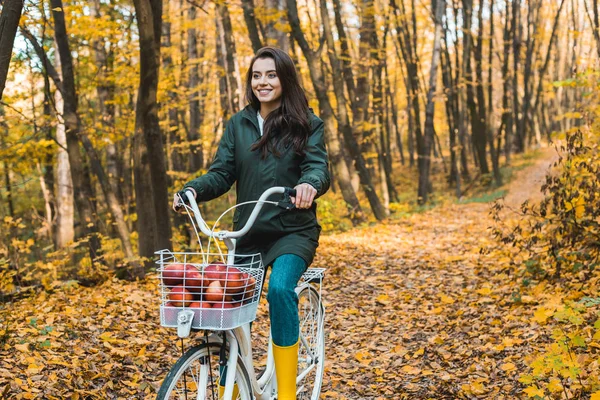 Chica feliz montando en bicicleta con cesta llena de manzanas en el bosque otoñal - foto de stock