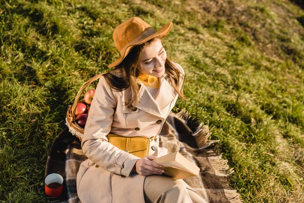 Vista de ángulo alto de la mujer elegante sonriente en el sombrero sentado en la manta y libro de lectura al aire libre - foto de stock