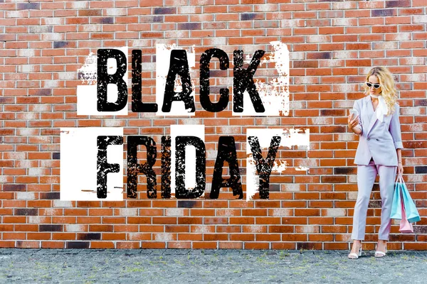 Stylish young woman with shopping bags using smartphone while standing in front of brick wall, black friday banner — Stock Photo