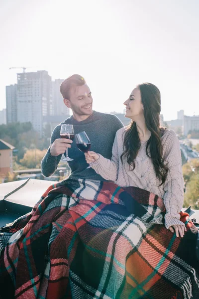 Happy young couple covering in plaid and clinking glasses of red wine on rooftop — Stock Photo