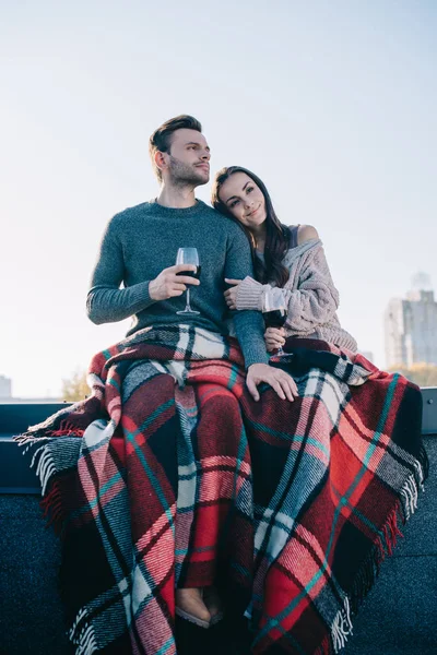 Bottom view of beautiful young couple covering in plaid and drinking red wine on rooftop — Stock Photo