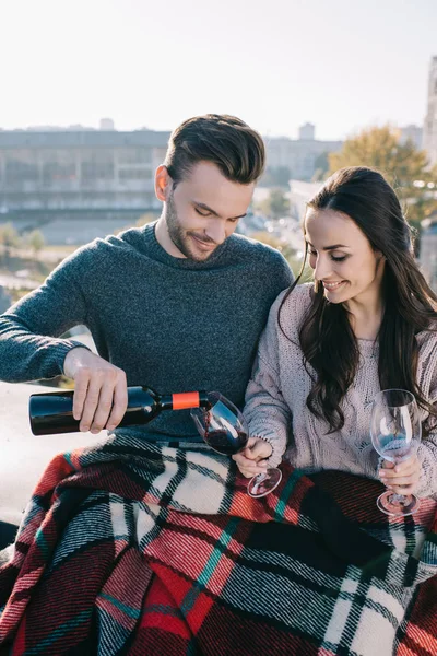Happy young couple covering in plaid on rooftop and pouting red wine into glasses — Stock Photo