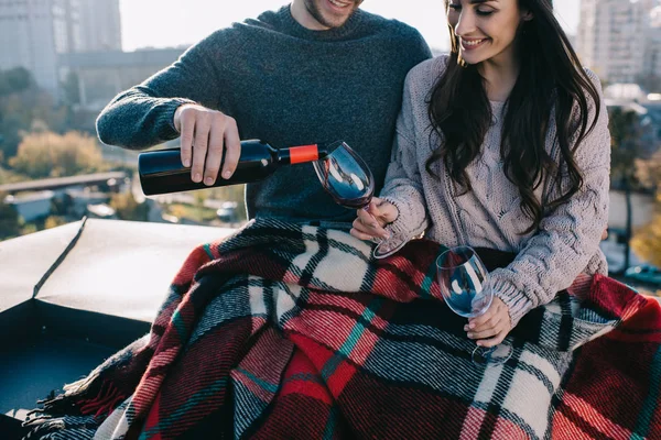 Tiro cortado de feliz jovem casal cobrindo em xadrez no telhado e derramando vinho tinto em copos — Fotografia de Stock