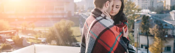 Panoramic shot of beautiful young couple covering in plaid and cuddling on rooftop — Stock Photo