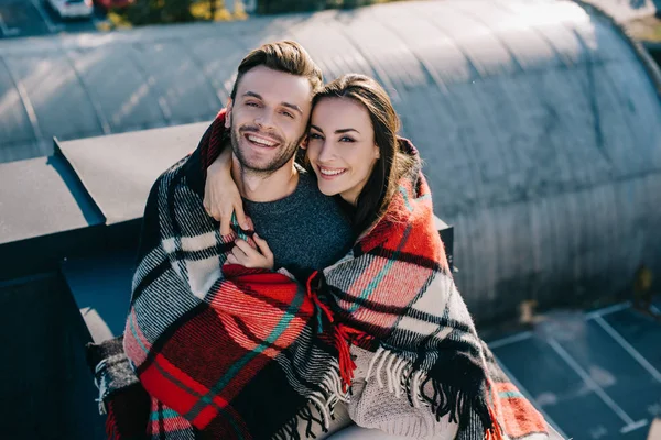 High angle view of beautiful young couple covered in plaid embracing and looking at camera on rooftop — Stock Photo