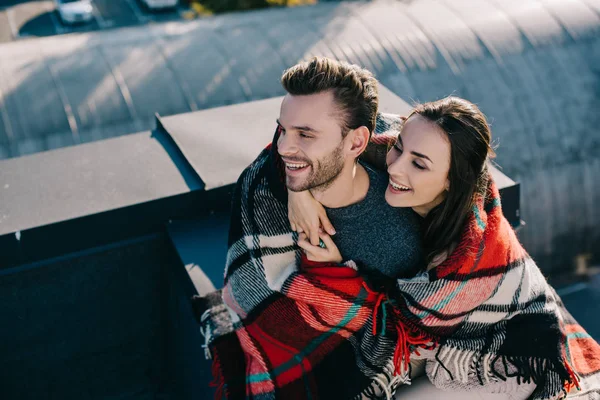 Vista de ángulo alto de la risa joven pareja cubierta de cuadros sentados en la azotea juntos - foto de stock