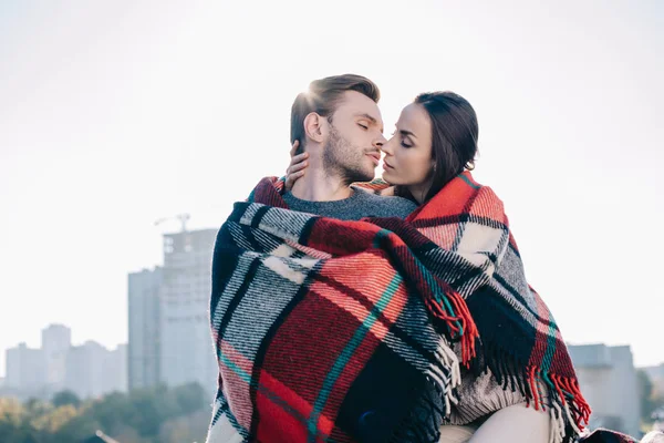 Schönes junges Paar, das sich in Plaid hüllt und sich küsst, während die Sonne dahinter scheint — Stockfoto