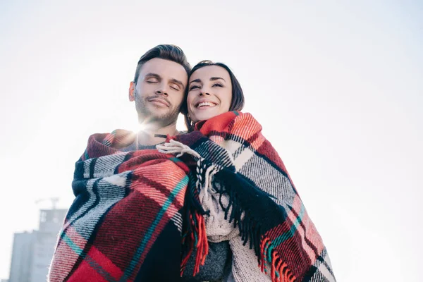 Bottom view of beautiful young couple covering in plaid and embracing with sun shining behind — Stock Photo