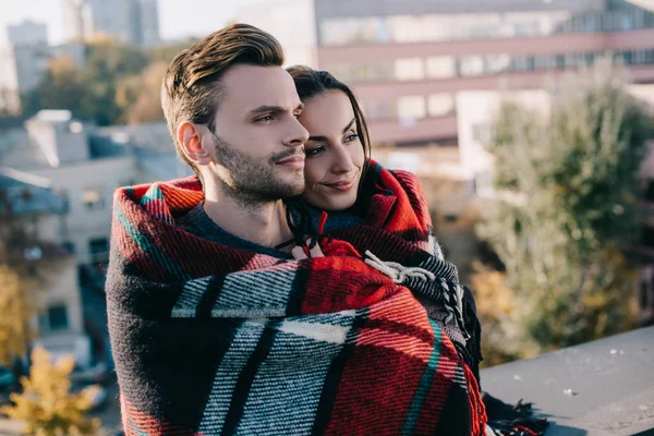 Lächelndes junges Paar, das sich in Karo hüllt und mit verschwommener Stadt im Hintergrund wegsieht — Stockfoto