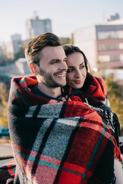 Glückliches junges Paar, das sich in Karo hüllt und mit verschwommener Stadt im Hintergrund wegsieht — Stockfoto