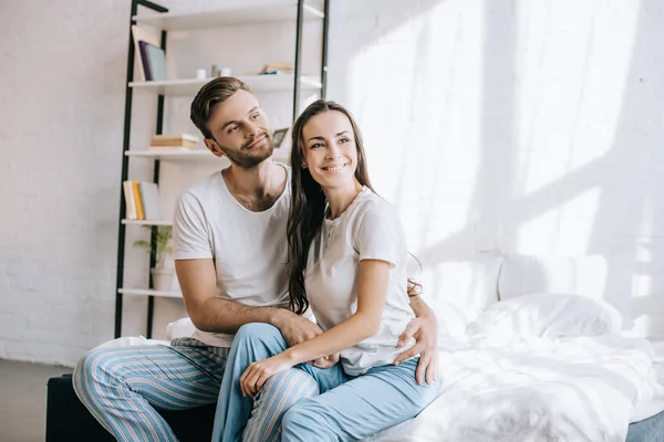 Feliz joven pareja en pijama sentado en la cama por la mañana y mirando hacia otro lado - foto de stock