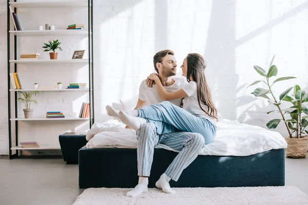 Schönes junges Paar im Schlafanzug kuschelt, während es morgens auf dem Bett sitzt — Stockfoto