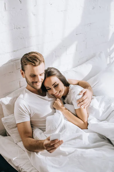 Vue grand angle de beau jeune couple se détendre au lit le matin et en utilisant smartphone — Photo de stock