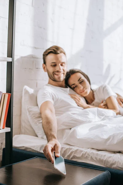 Jovem feliz relaxando na cama com a namorada e alcançando o smartphone — Fotografia de Stock