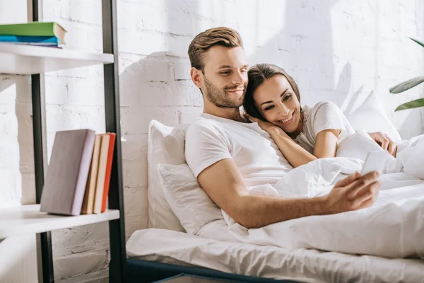 Belo jovem casal relaxante na cama de manhã e usando smartphone — Fotografia de Stock