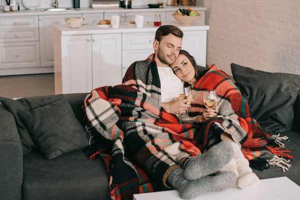 Beautiful young couple with glasses of white wine relaxing on couch under plaid — Stock Photo