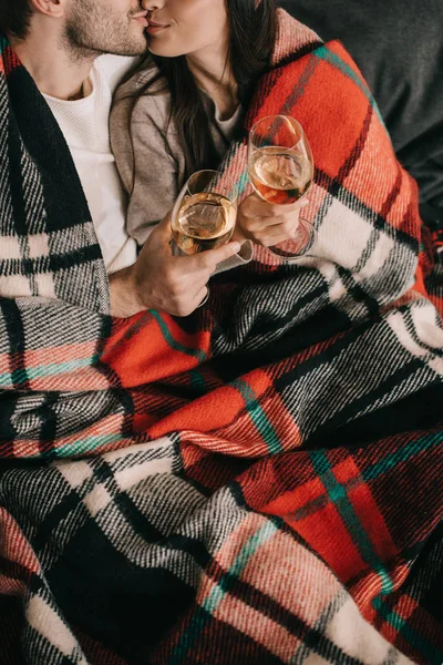 Cropped shot of young couple with glasses of white wine relaxing and kissing on couch under plaid — Stock Photo