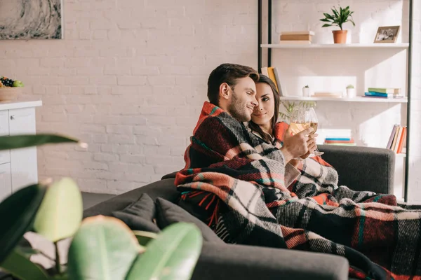 Happy young couple with glasses of white wine relaxing on couch under plaid — Stock Photo