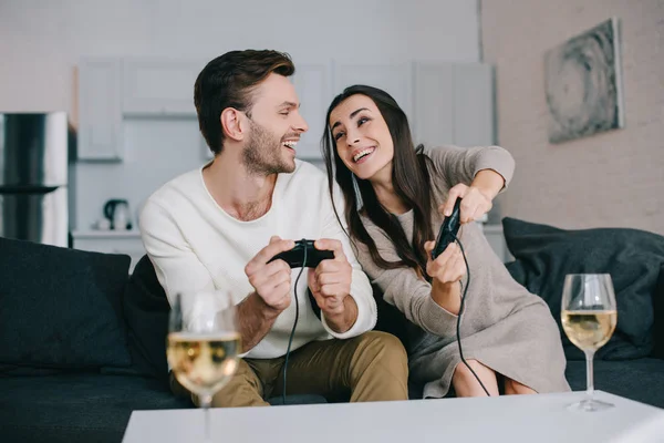 Счастливая молодая пара с бокалами вина, играющая в ретро видео игру на диване дома — стоковое фото