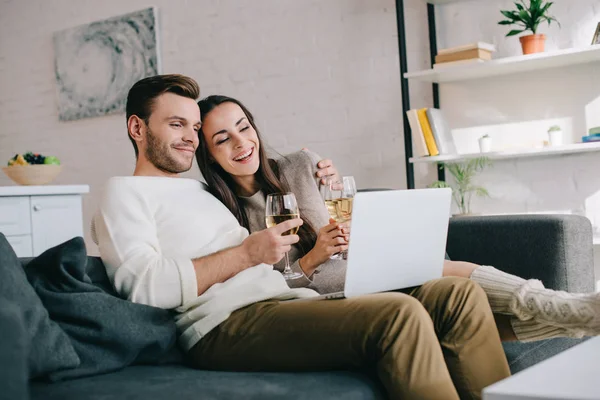 Rire jeune couple en utilisant un ordinateur portable et boire du vin ensemble sur le canapé à la maison — Photo de stock