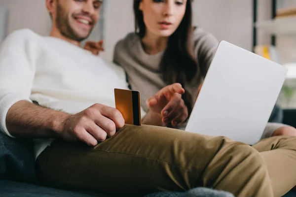 Bella giovane coppia che fa e-shopping con carta di credito dorata e laptop sul divano di casa — Foto stock