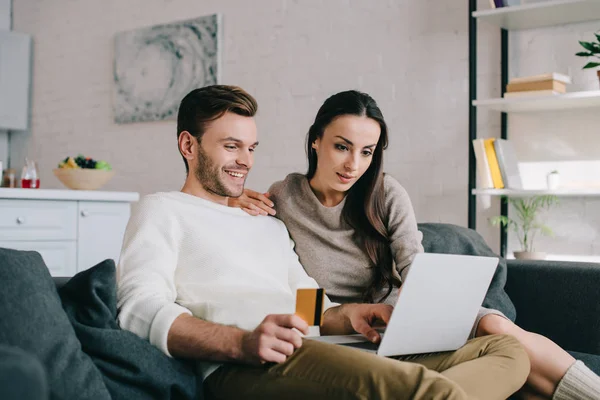 Sonriente joven pareja haciendo e-shopping con portátil en el sofá en casa - foto de stock