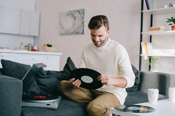 Schöner junger Mann hält Schallplatte für Plattenspieler, während er auf der Couch sitzt — Stockfoto