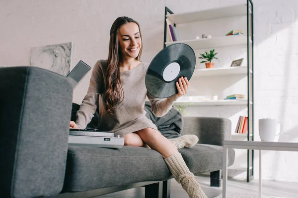 Happy young woman listening music with vinyl record player on couch at home — Stock Photo