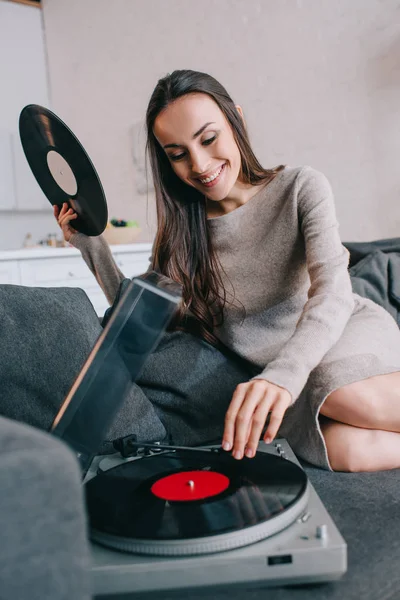 Sorridente giovane donna che ascolta musica con giradischi in vinile sul divano di casa — Foto stock