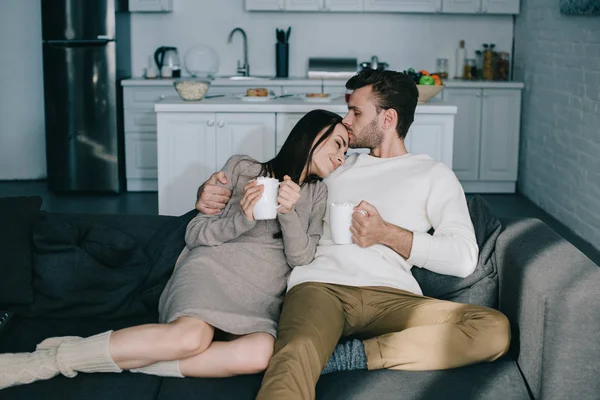 Belo jovem casal com xícaras de cacau com marshmallow abraçando no sofá em casa — Fotografia de Stock