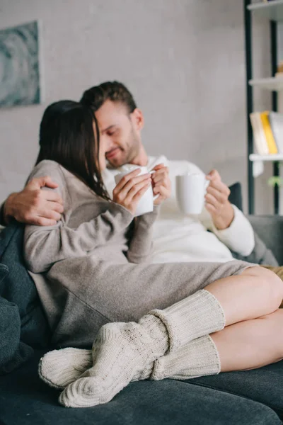 Feliz jovem casal com xícaras de cacau com marshmallow abraçando no sofá em casa — Fotografia de Stock