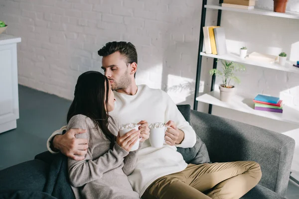 Hermosa pareja joven con tazas de cacao con malvavisco sentado en el sofá en casa - foto de stock