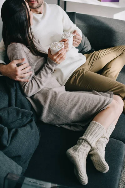 Tiro recortado de pareja con tazas de cacao en calcetines de lana cálidos relajándose en el sofá en casa - foto de stock