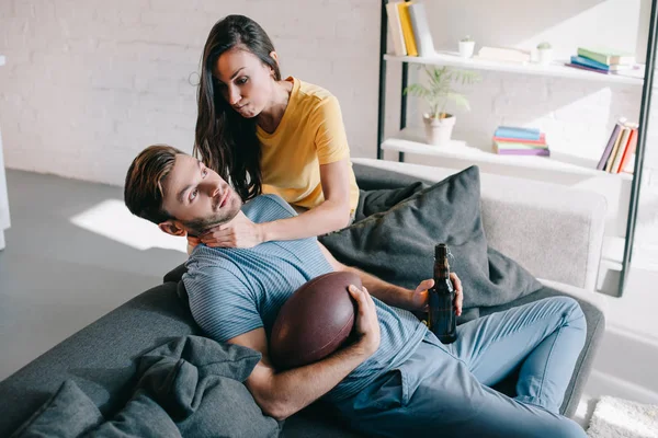 Сердитая женщина душит своего мужа, когда он смотрит американский футбол по телевизору дома — стоковое фото
