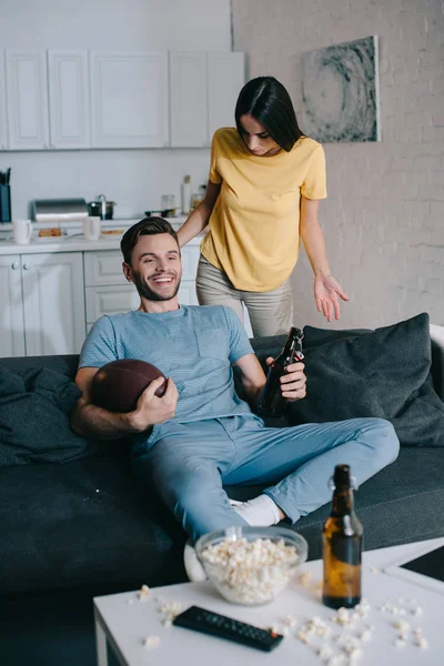 Сердита жінка кричить на чоловіка, коли він дивиться американський футбол на телебаченні вдома — стокове фото