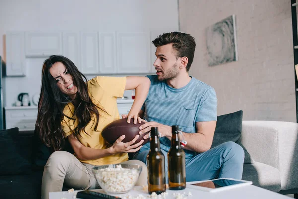 Сердитая молодая женщина борется за американский футбольный мяч с мужем во время матча по телевизору дома — стоковое фото