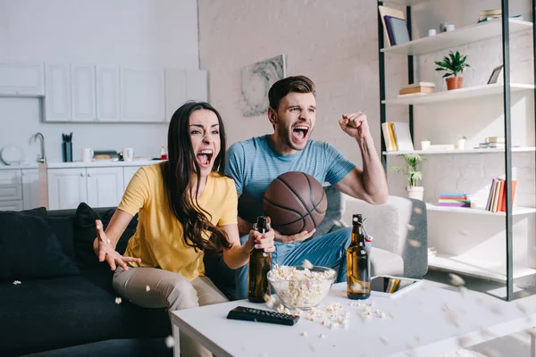 Expressivo jovem casal com cerveja torcendo para o jogo de basquete em casa — Fotografia de Stock