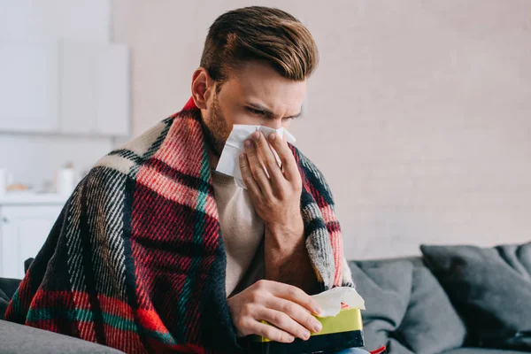 Sick young man sneezing with paper napkins sitting on couch — Stock Photo