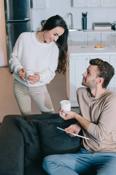 Lächelndes junges Paar mit Tassen Kakao und Tablette entspannt zu Hause — Stockfoto