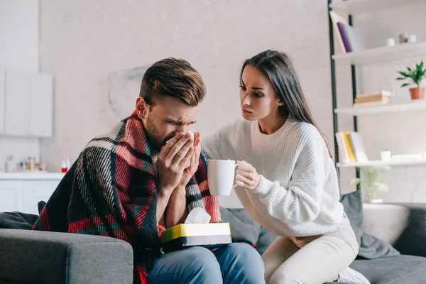 Hermosa mujer joven sosteniendo taza de bebida caliente para novio enfermo mientras estornuda - foto de stock